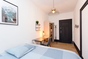 Chambre minimaliste avec lit blanc au premier plan, bureau noir avec chaise grise, murs blancs et portes noires, parquet clair
