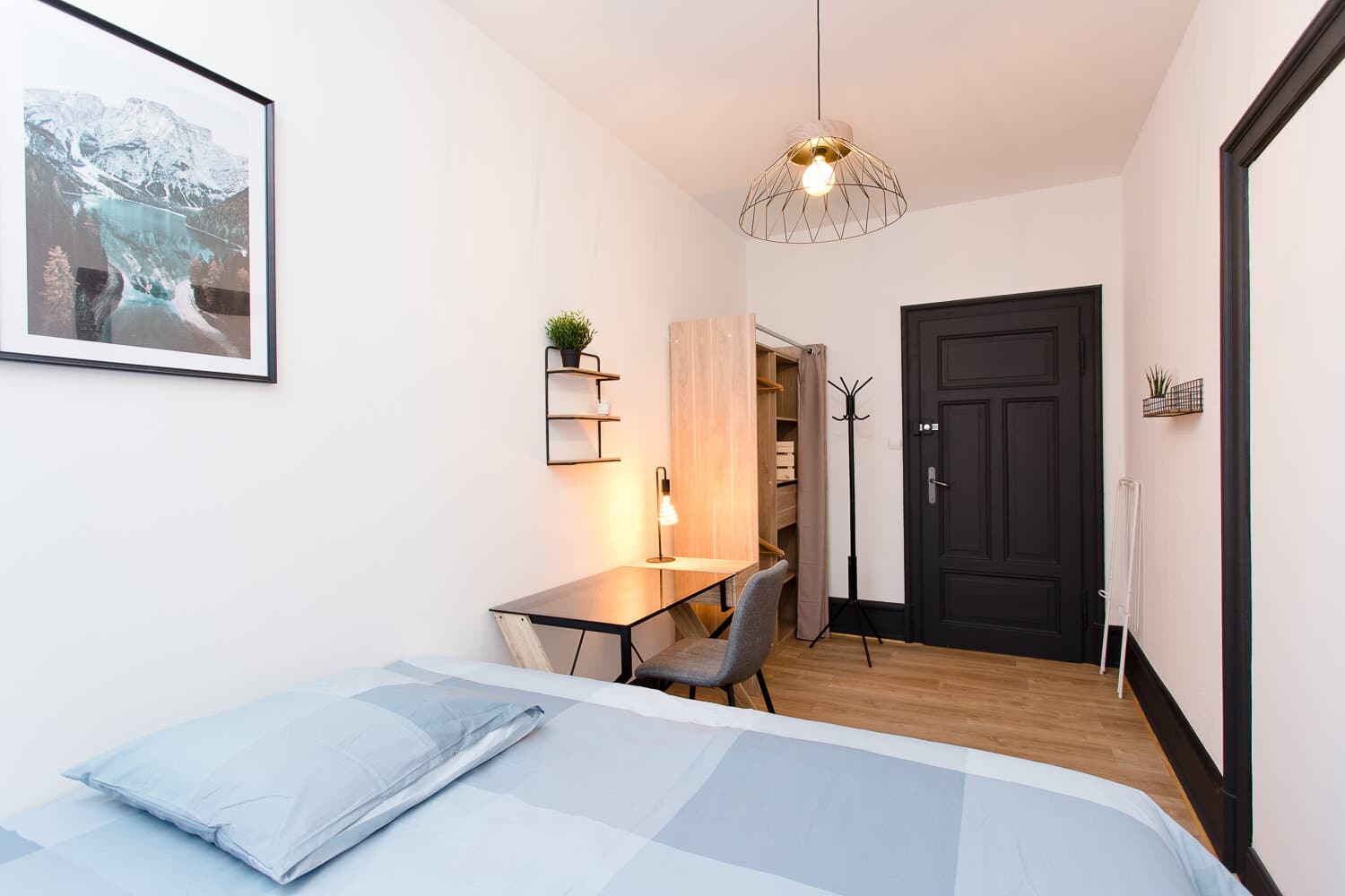 Chambre spacieuse avec lit blanc au premier plan, bureau noir et chaise grise face à la fenêtre, murs clairs et porte noire.