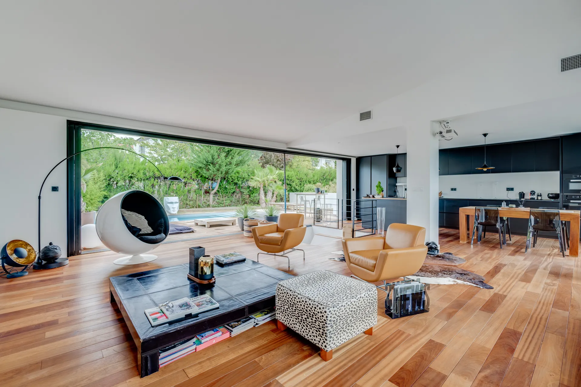 Salon spacieux avec fauteuils en cuir camel, table basse noire et vue sur terrasse boisée, cuisine ouverte au fond