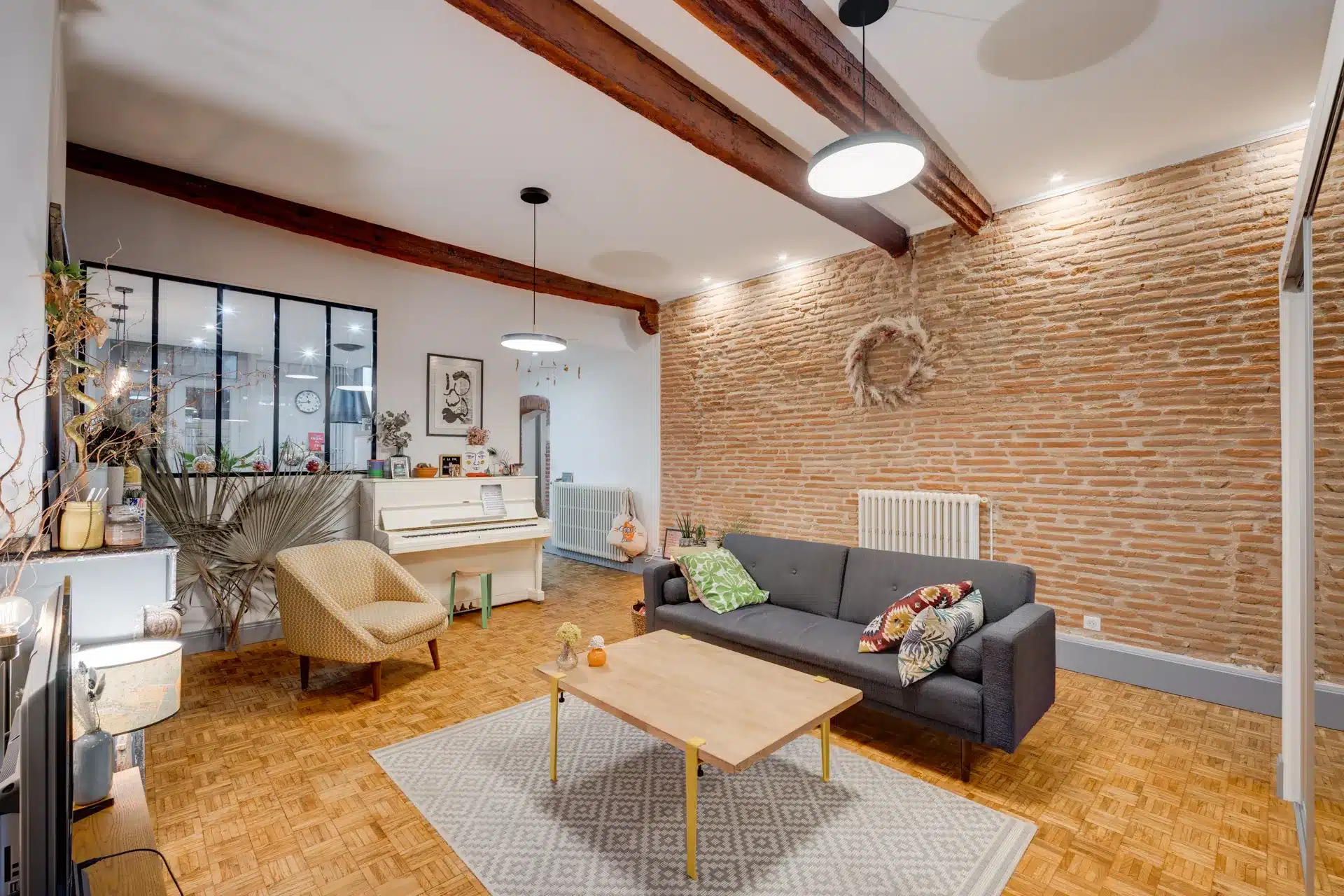 Loft spacieux avec murs en brique rouge, poutres bois au plafond, canapé gris et fauteuil jaune, sol parquet.