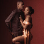 Couple posant ensemble sur fond rouge, femme en robe or et homme en chemise noire