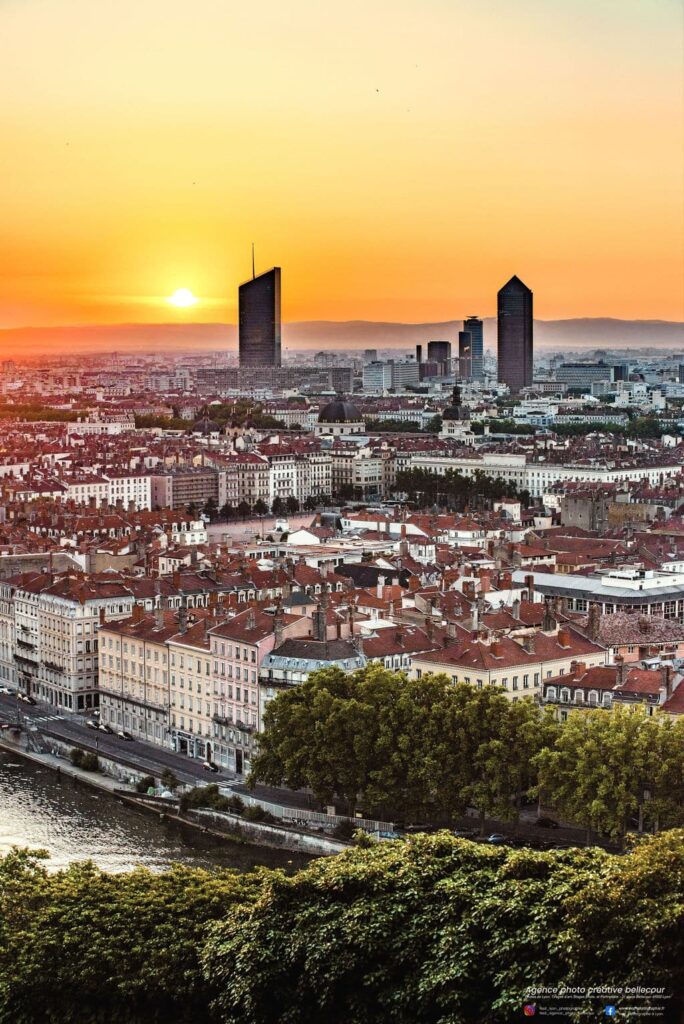 Vue matinale du lever de soleil sur la skyline de Lyon en 2022, prise depuis le Jardin des Curiosités, avec des teintes rosées et dorées éclairant le ciel et la ville.
