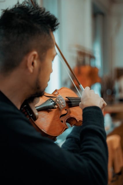 Musicien en pull noir joue du violon, vue de profil, dans une salle avec d'autres musiciens en arrière-plan