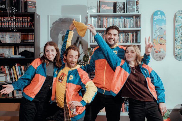 Quatre personnes en vestes de sport bleu et orange sourient et posent ensemble dans une pièce remplie de livres et d'affiches de cinéma