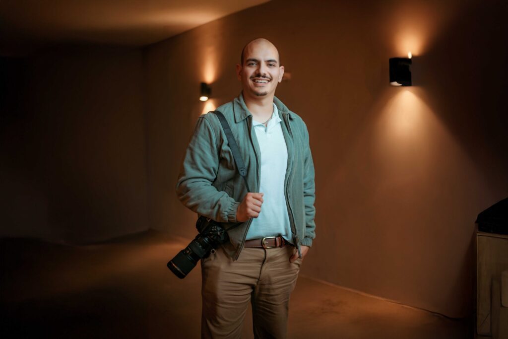 Homme souriant tenant un appareil photo reflex en intérieur, vêtu d'une veste grise et chemise blanche