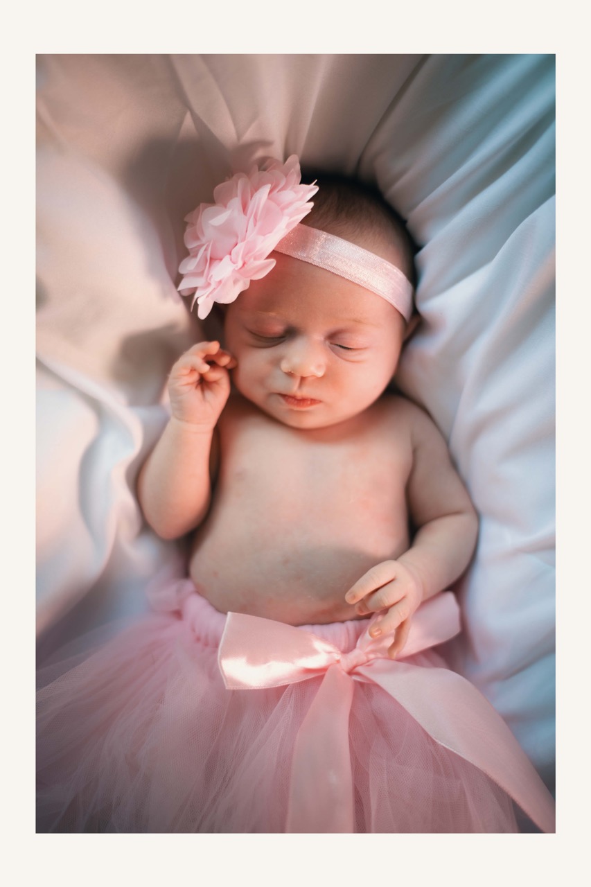 Bébé en robe rose avec une fleur dans les cheveux, paisiblement allongé sur un fond doux.