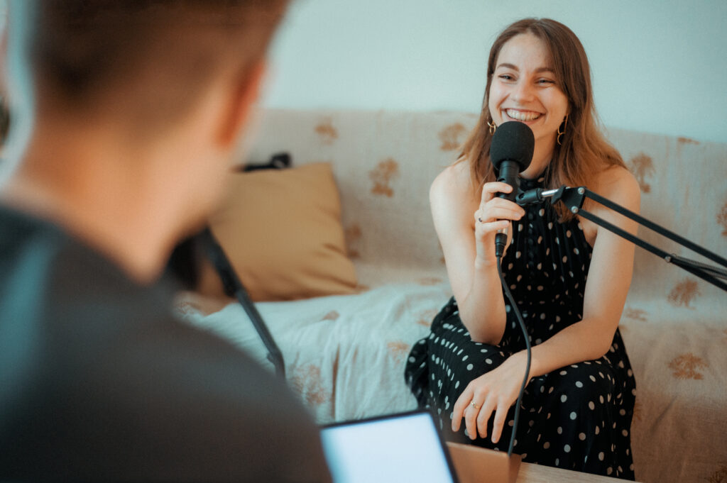 Femme en robe noire à pois sourit en tenant un microphone face à un homme de dos assis sur un canapé beige