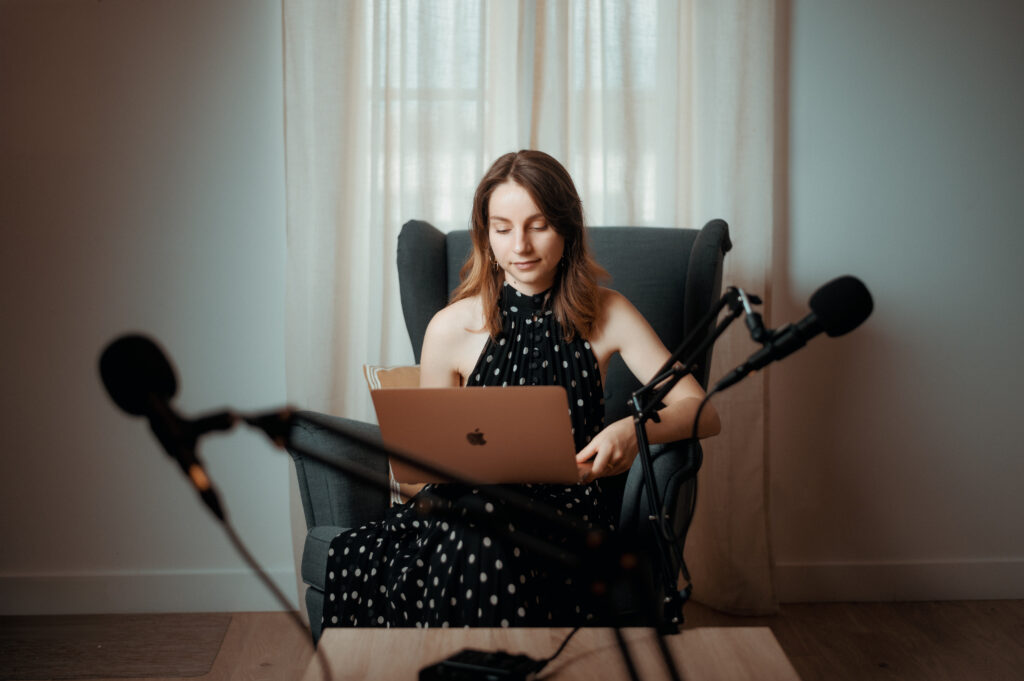 Femme assise dans un fauteuil gris, robe noire à pois, tenant un laptop et parlant dans un microphone, lumière naturelle par la fenêtre