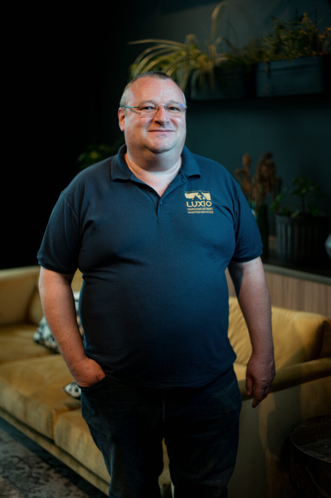 Homme souriant en polo bleu marine debout dans un salon avec canapé beige et plantes vertes en arrière-plan