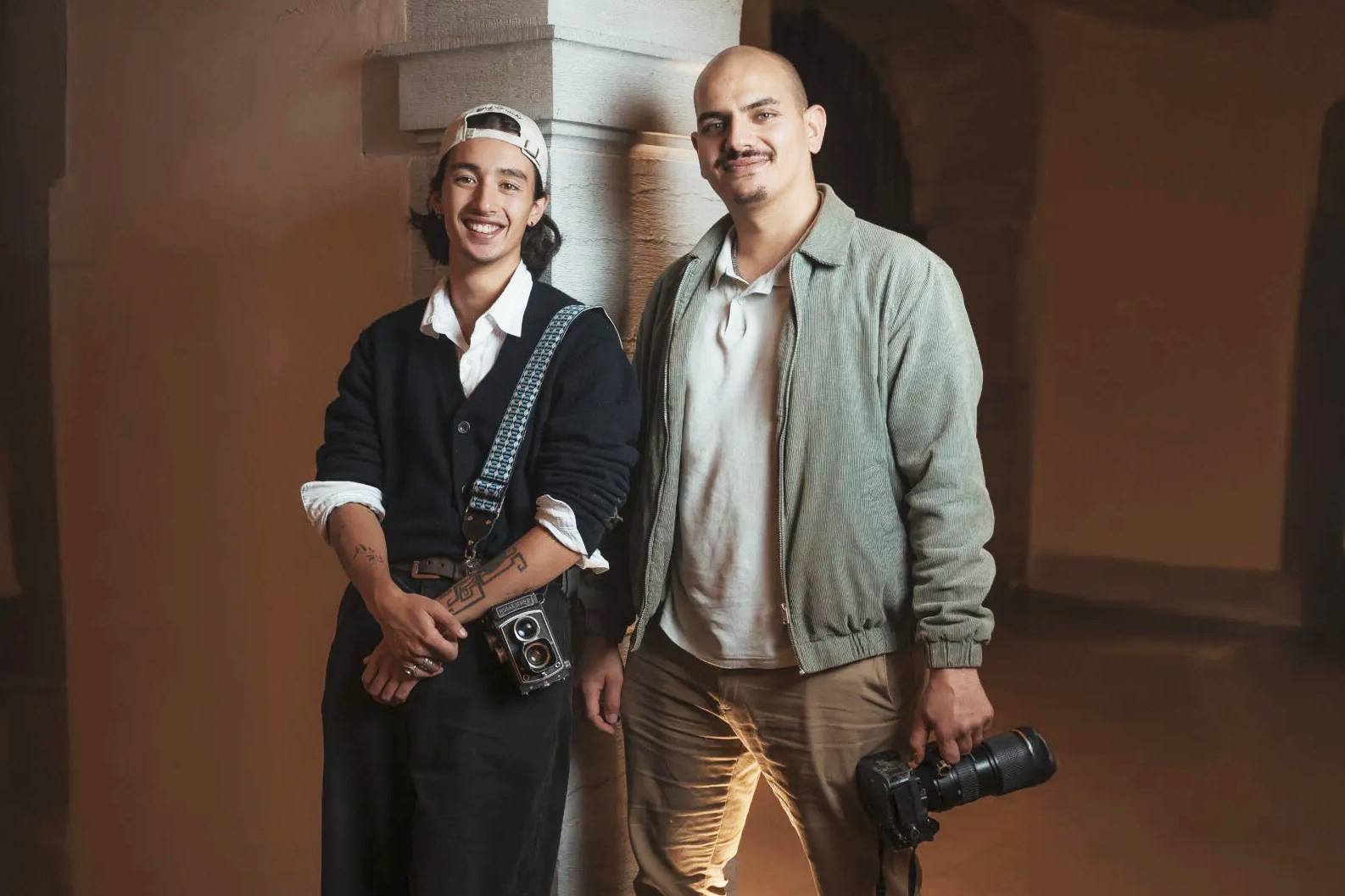 Deux hommes souriants tenant des appareils photo dans un studio aux tonalités chaudes. Nicolas et Kaito de Ned Photographie