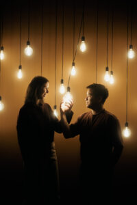 Couple se tenant la main sous ampoules LED suspendues, lumière dorée chaude, fond sombre