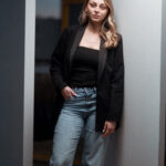 Femme en blazer noir et jean bleu posée entre deux murs blancs, regardant vers le bas