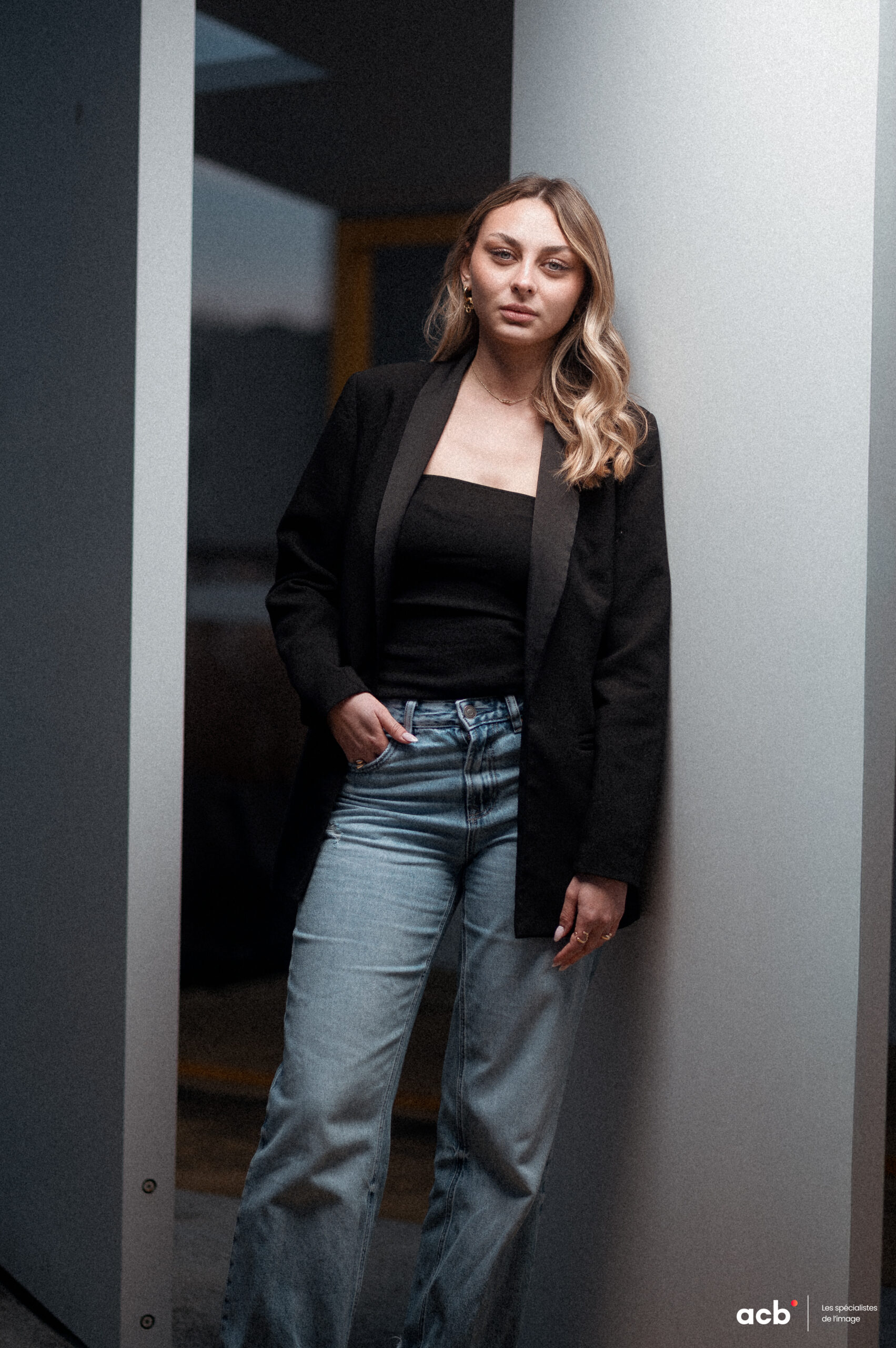 Femme en blazer noir et jean bleu posée entre deux murs blancs, regardant vers le bas