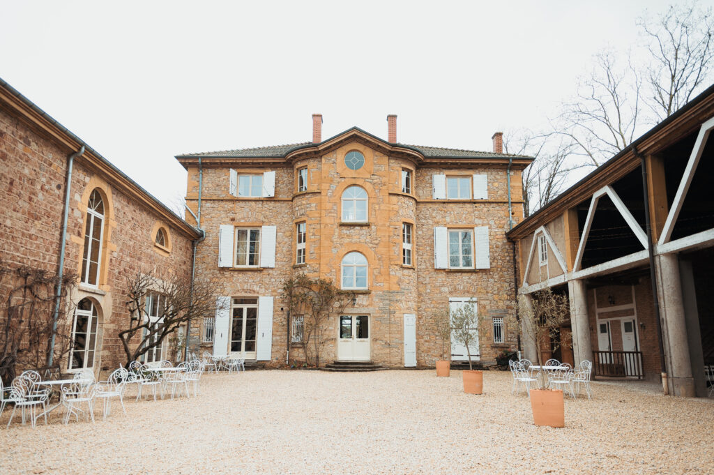 Cour intérieure d'un château en pierre avec chaises blanches disposées face à la façade, bâtisses en briques et ciel gris
