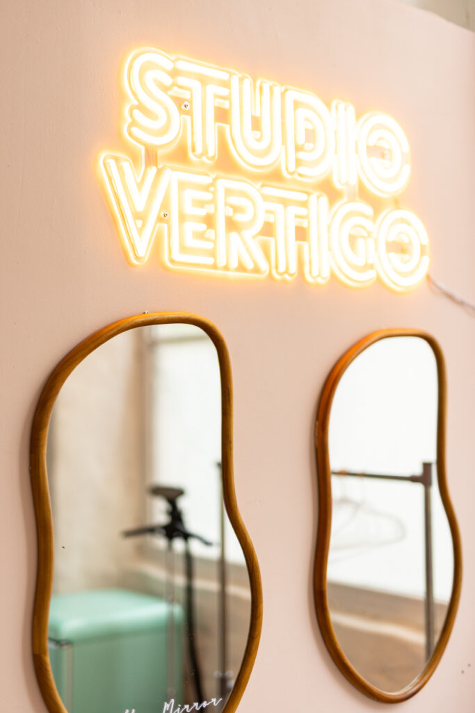 Enseigne lumineuse jaune Studio Vertigo au-dessus de deux miroirs ovales aux cadres bois