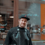 Homme en casquette noire et tablier gris sourit devant la vitrine de son atelier en ville