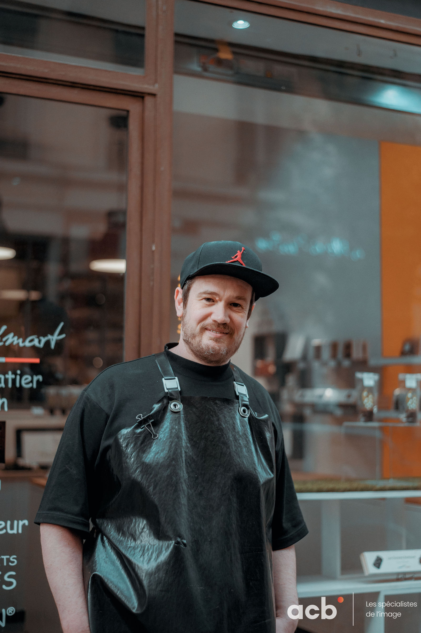 Homme en casquette noire et tablier gris sourit devant la vitrine de son atelier en ville