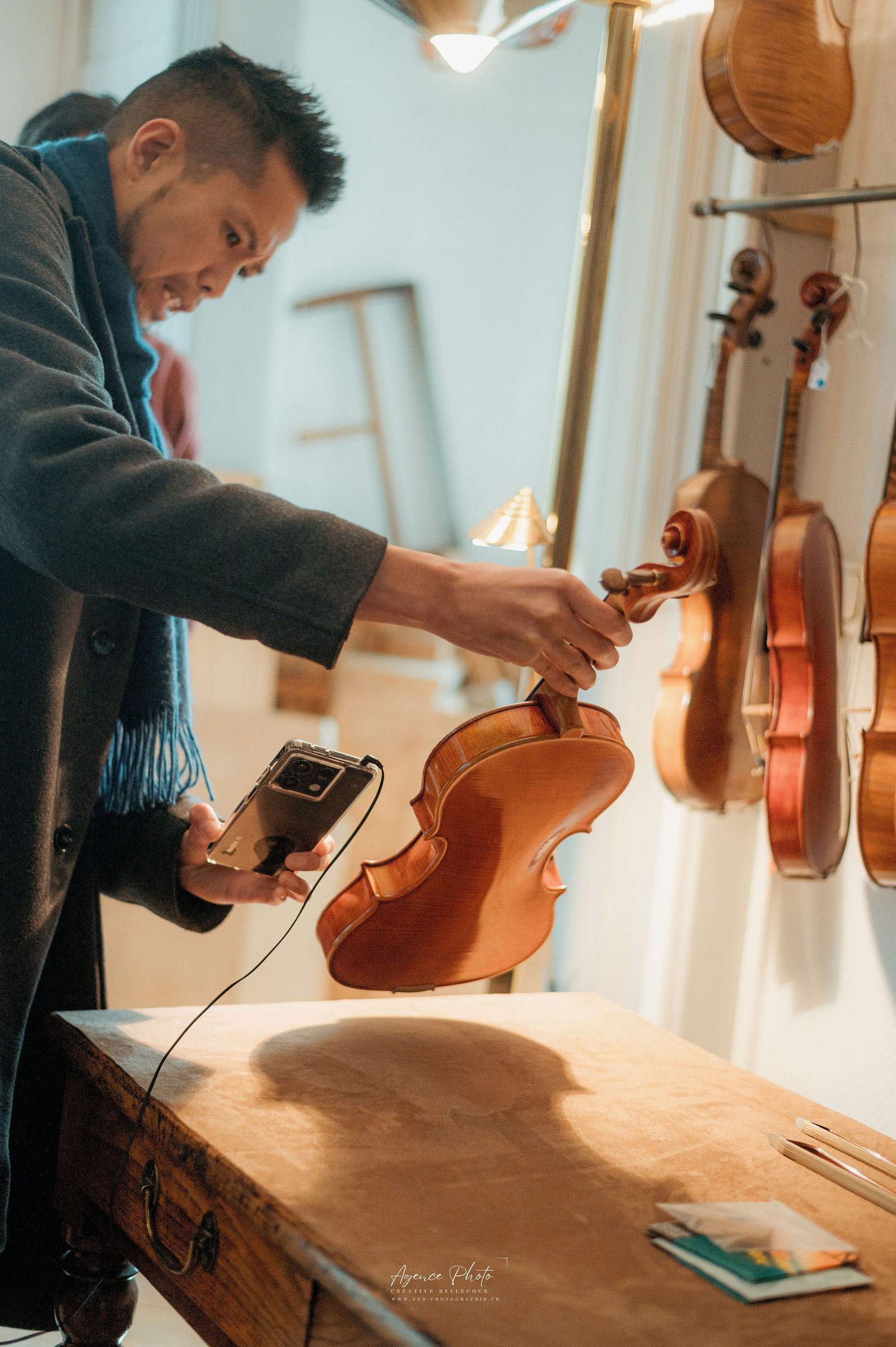 Homme examine un violon en bois avec son téléphone dans un atelier, plusieurs instruments suspendus au mur