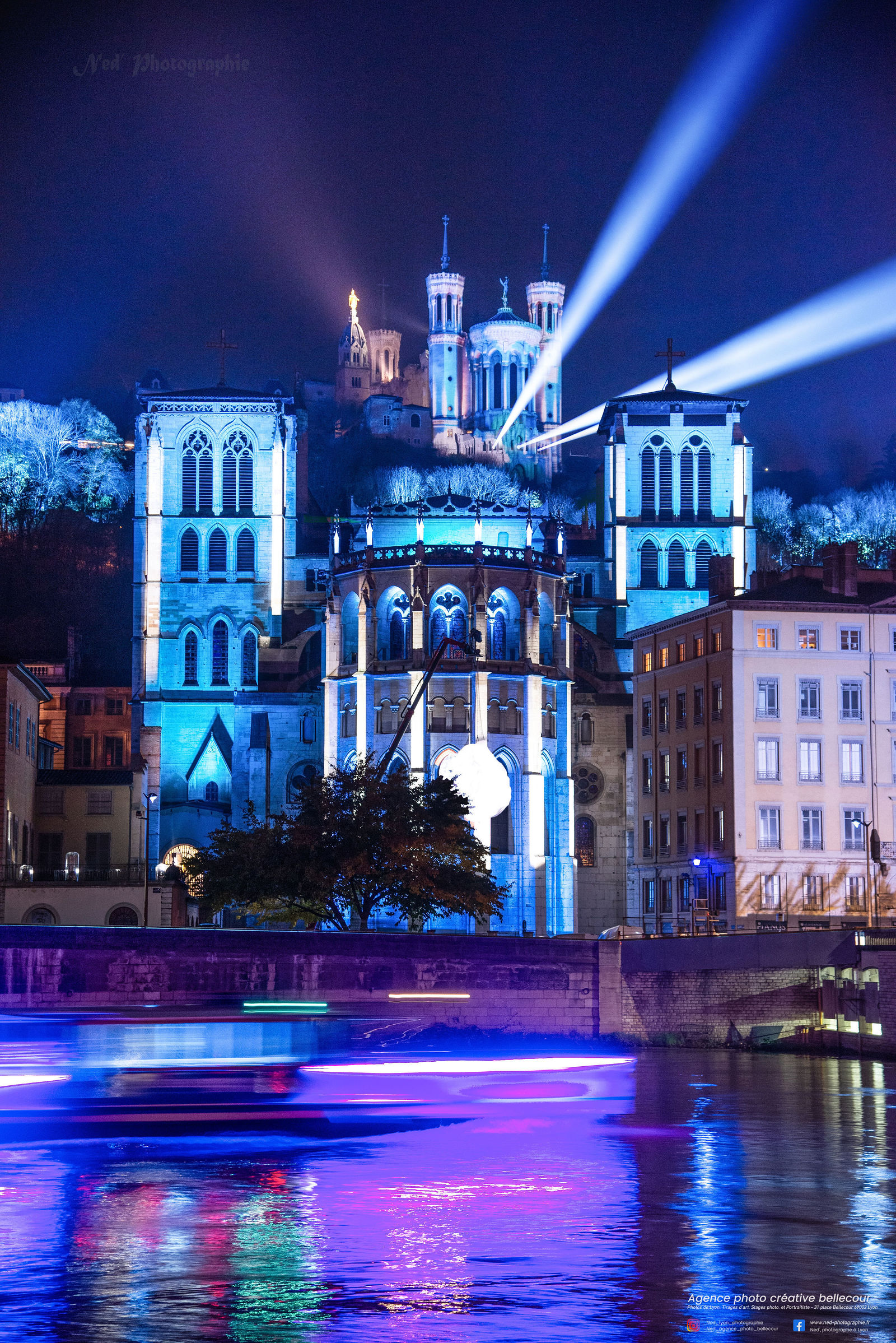 Basilique de Fourvière éclairée en bleu et violet la nuit, avec la Saône en premier plan reflétant les lumières colorées