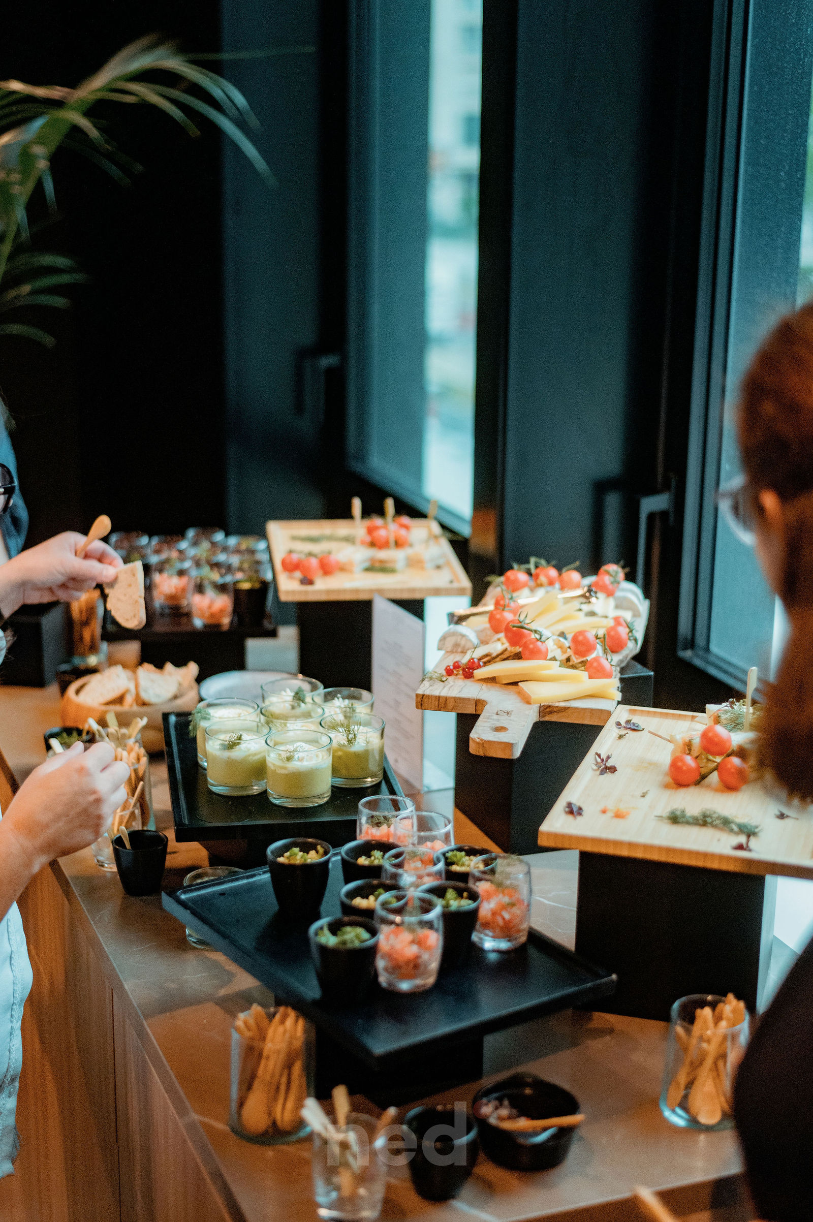 Deux personnes dégustent des canapés et des verres autour d'une table haute avec plateaux de fromages, tomates et créations culinaires