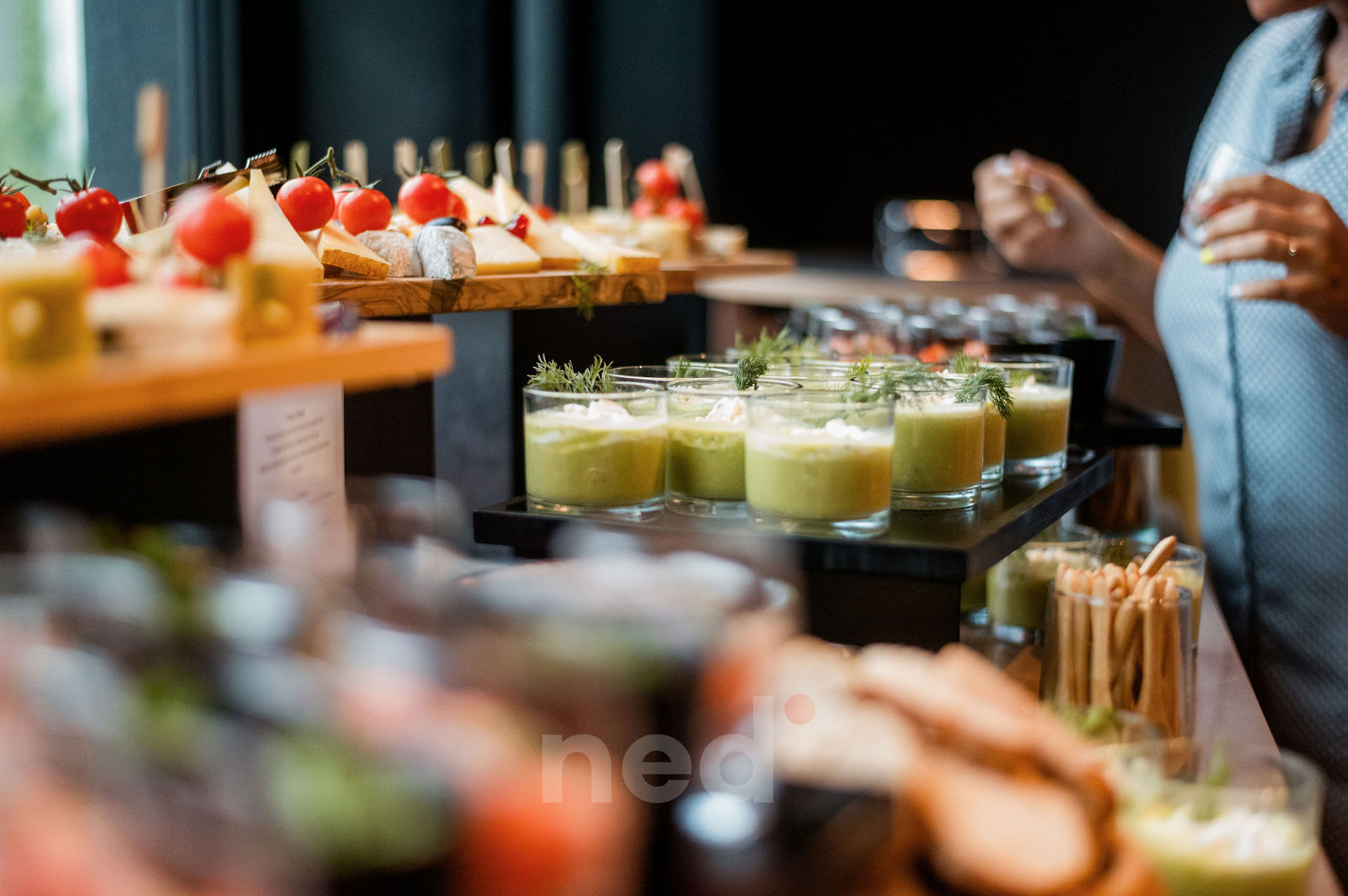 Table de réception avec verres de jus vert, canapés aux tomates cerises et fromage, femme enceinte en robe bleue à côté