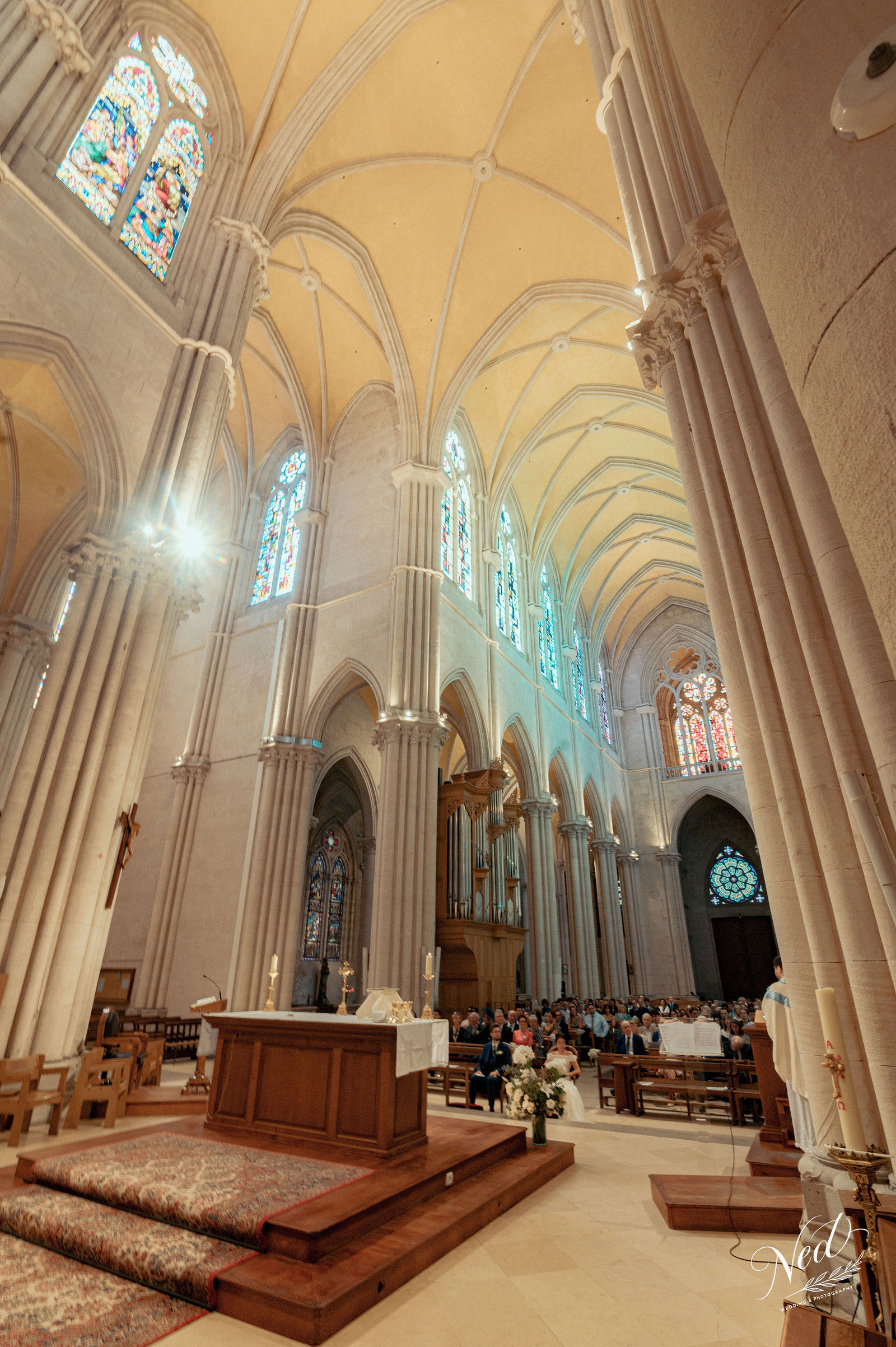 Nef de cathédrale gothique avec voûtes en ogives, invités assis dans les bancs, autel orné de fleurs blanches, lumière des vitraux bleus