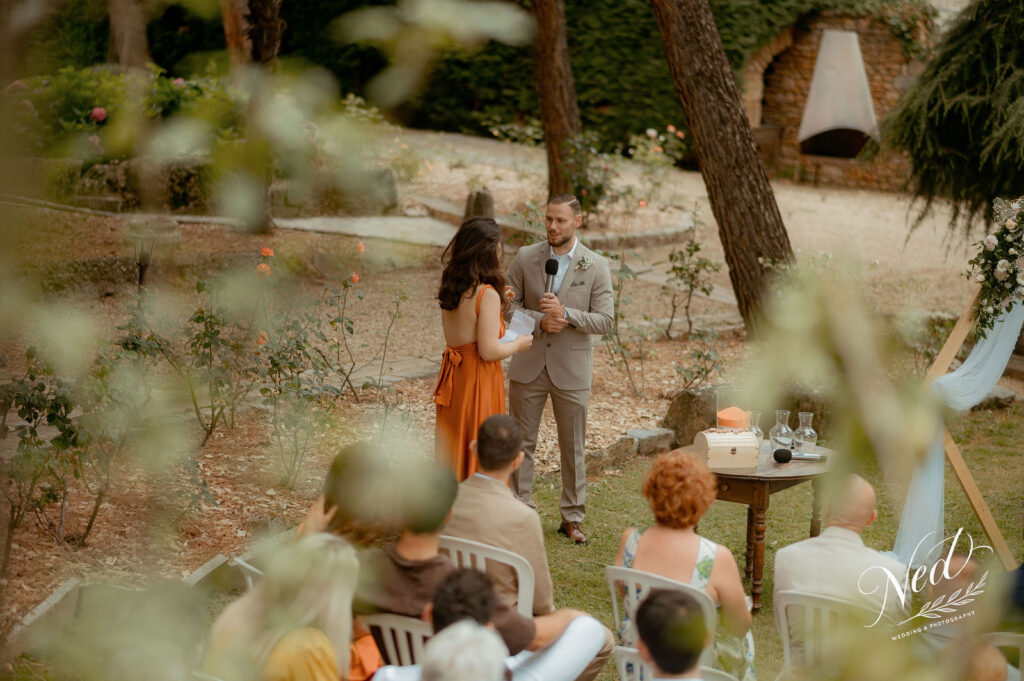 Homme en costume beige et femme en robe orange échangent leurs voeux devant des invités assis dans un jardin arboré