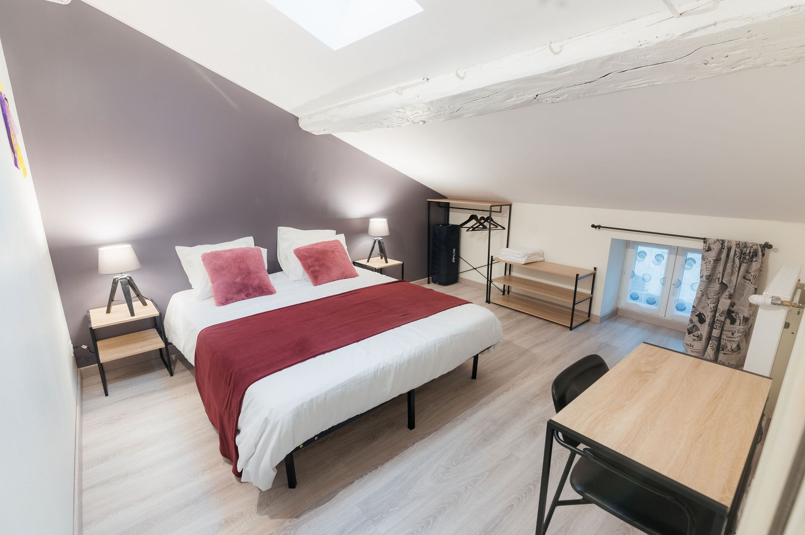 Chambre d'hôtel mansardée avec lit double blanc et chemin de lit rouge, deux lampes de chevet, murs gris clair et poutres blanches apparentes