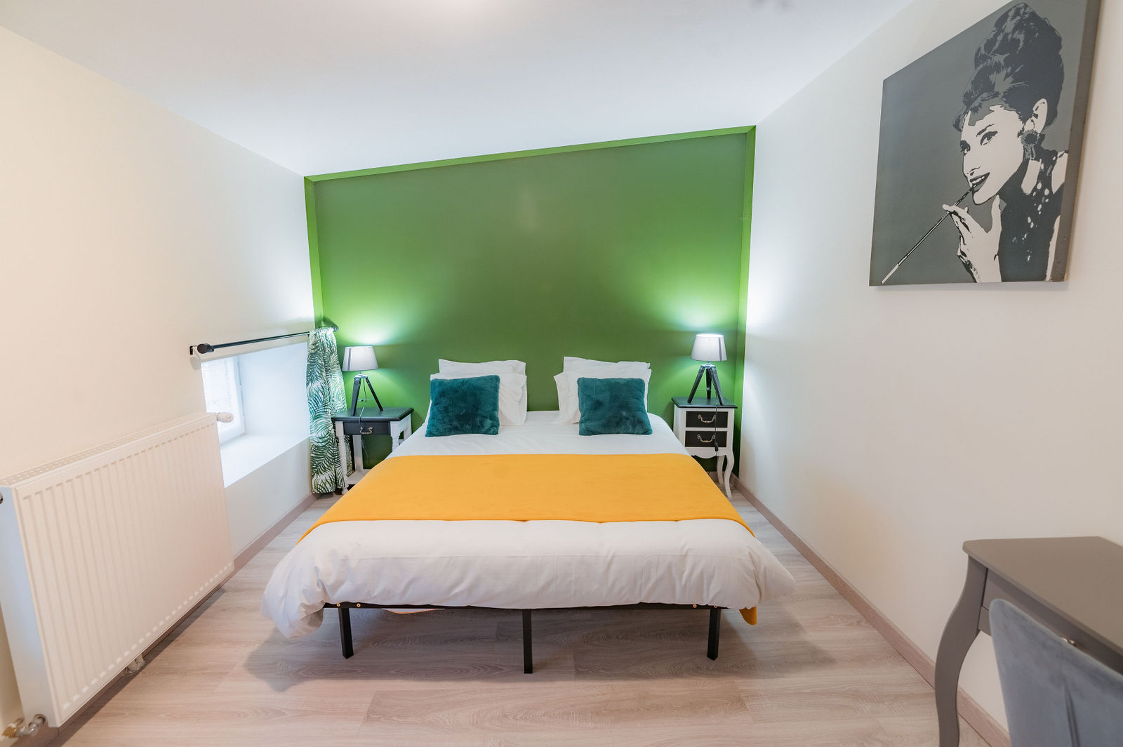 Chambre à coucher avec tête de lit verte, lit blanc et jaune, coussins bleu canard, lampes de chevet et portrait pop art au mur