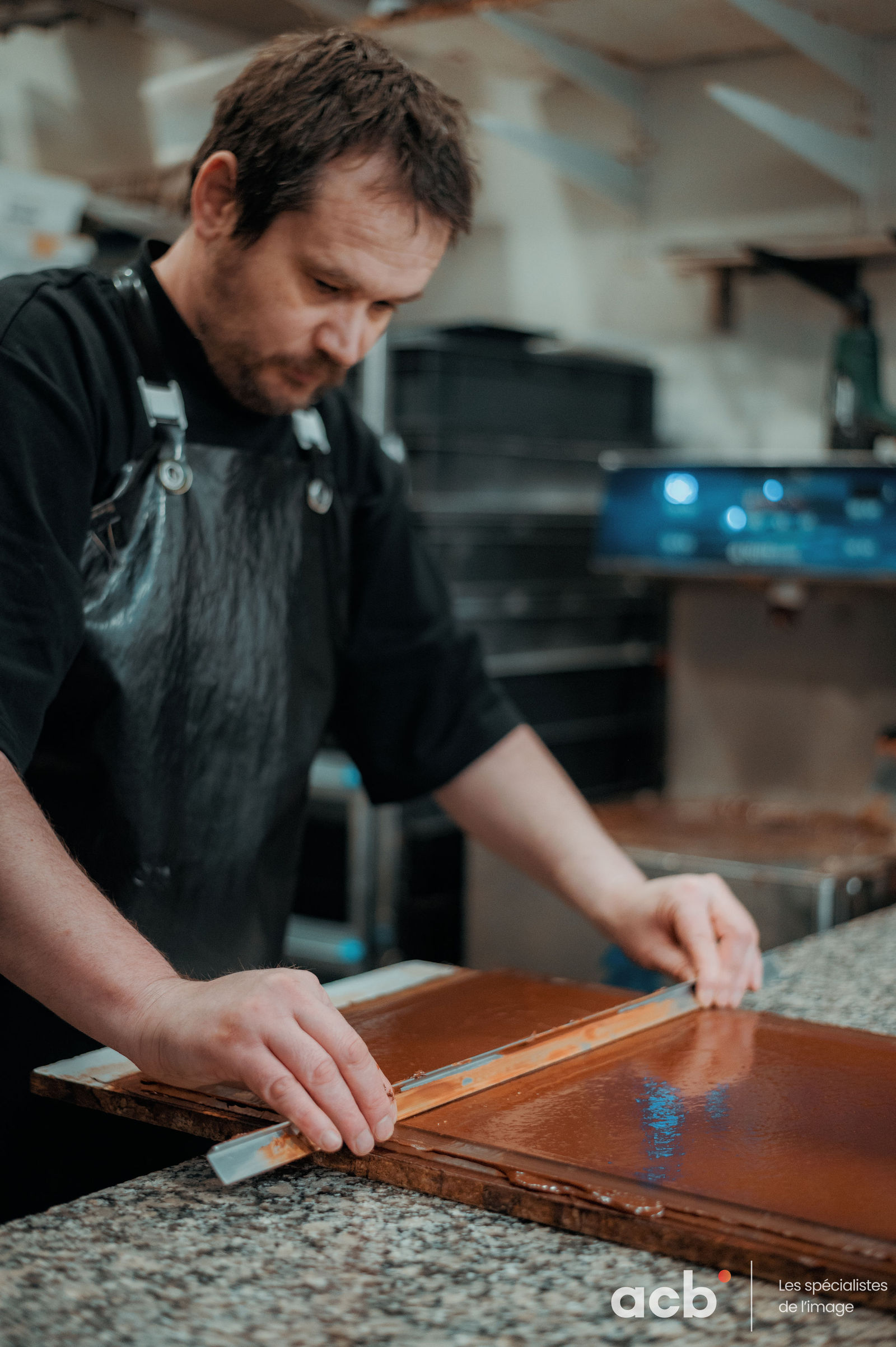 Chocolatier en tablier noir travaille une plaque de chocolat sur son plan de travail en atelier artisanal