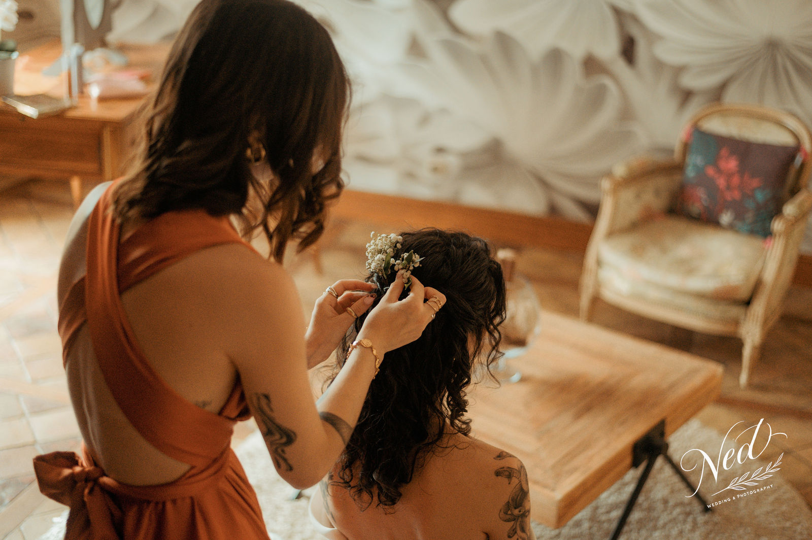 Femme en robe terracotta ajuste les fleurs blanches dans les cheveux d'une mariée aux cheveux noirs ondulés, intérieur élégant