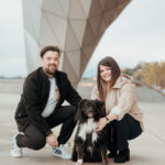 Couple souriant accroupi avec leur chien noir et blanc sous une structure architecturale moderne, berges du Rhône en arrière-plan