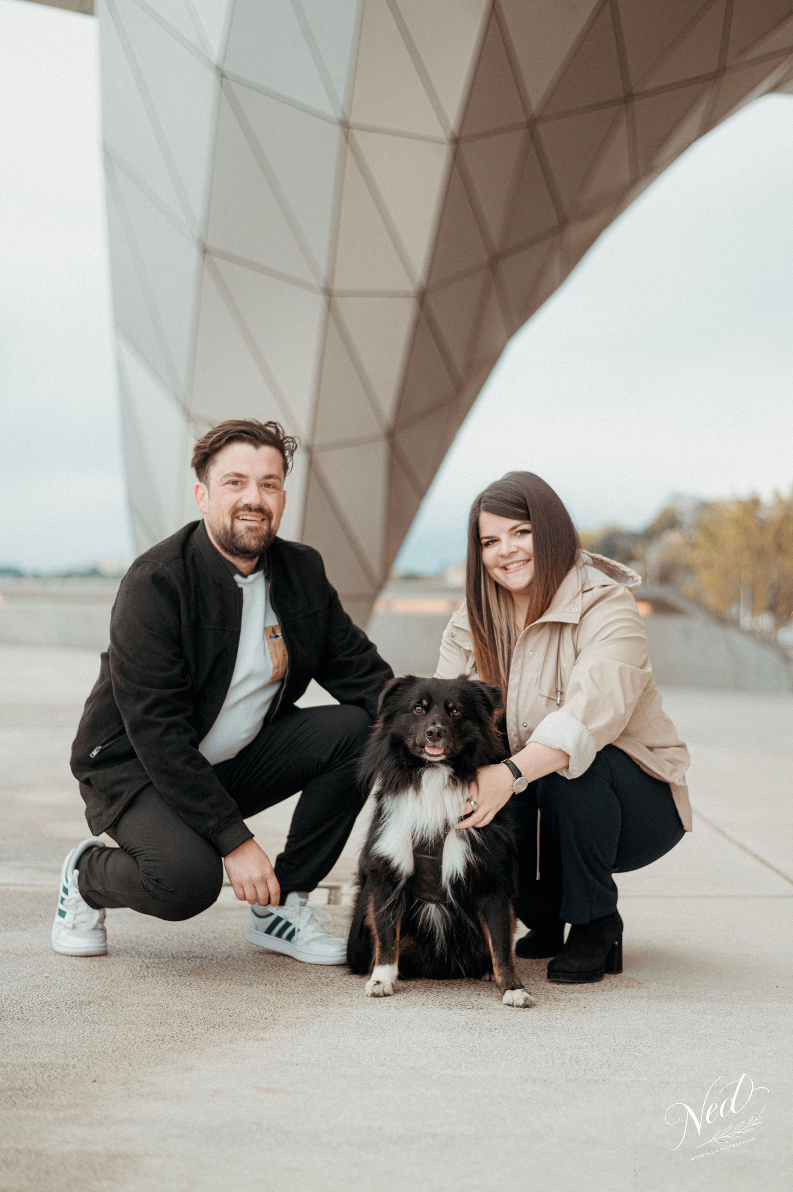 Couple souriant accroupi avec leur chien noir et blanc sous une structure architecturale moderne, berges du Rhône en arrière-plan