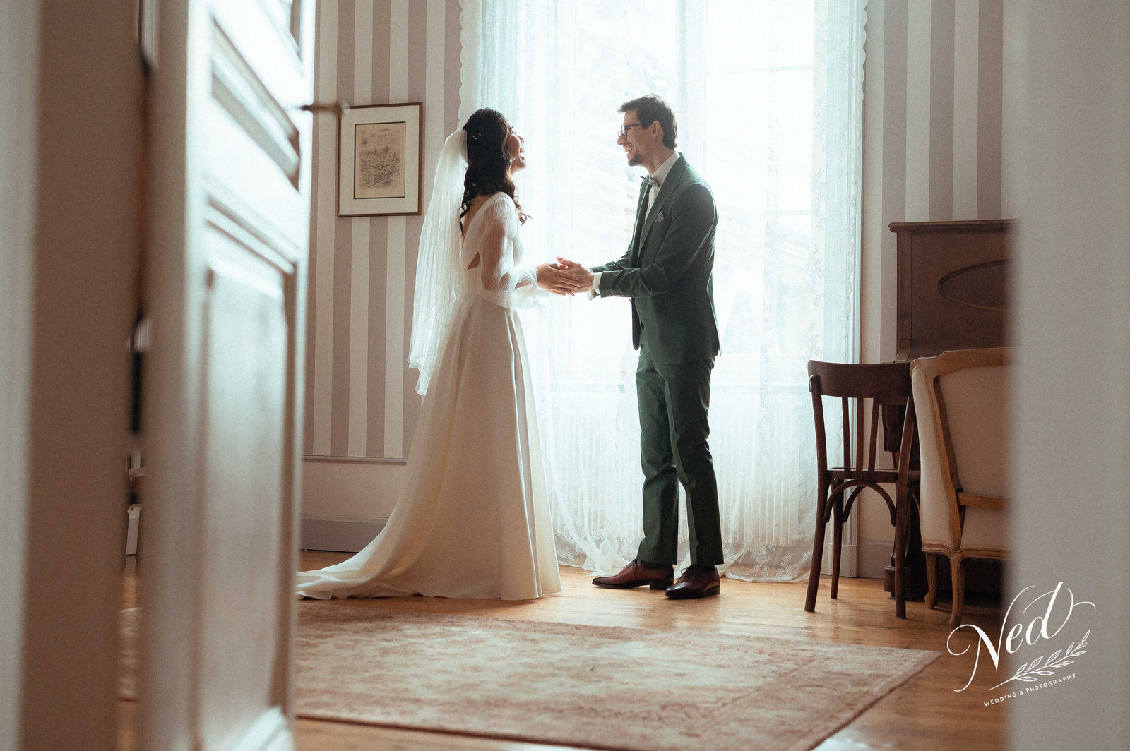 Mariée en robe blanche et voile face à son mari en costume gris, se tenant les mains dans une salle aux murs striés, lumière naturelle des fenêtres