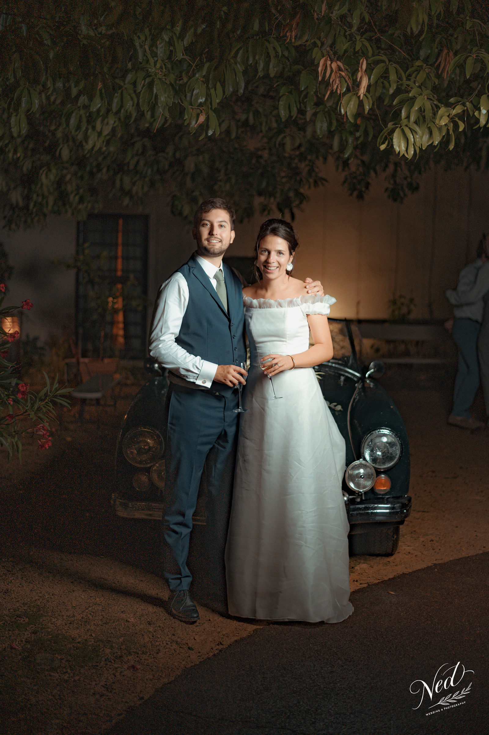 Mariée en robe blanche et marié en costume bleu posent enlacés devant un scooter noir, sous un feuillage, lumière chaude du soir