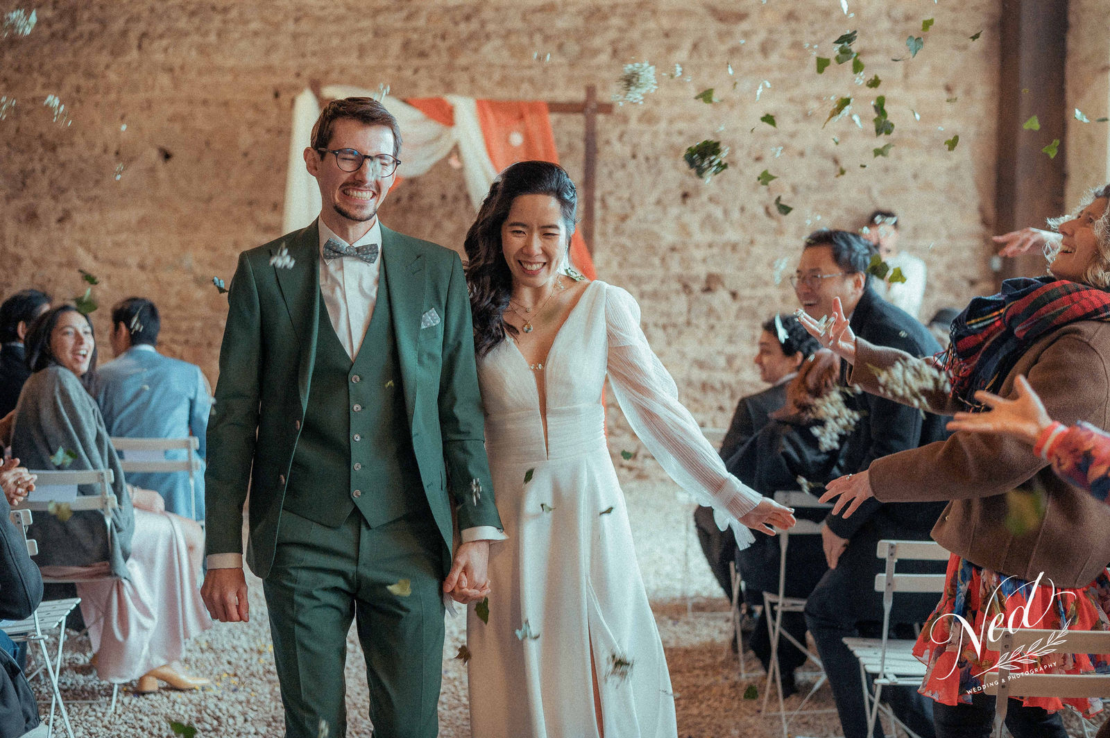 Jeune couple souriant sort de la cérémonie dans une salle en pierre, confettis verts et blancs volent autour d'eux, invités applaudissent