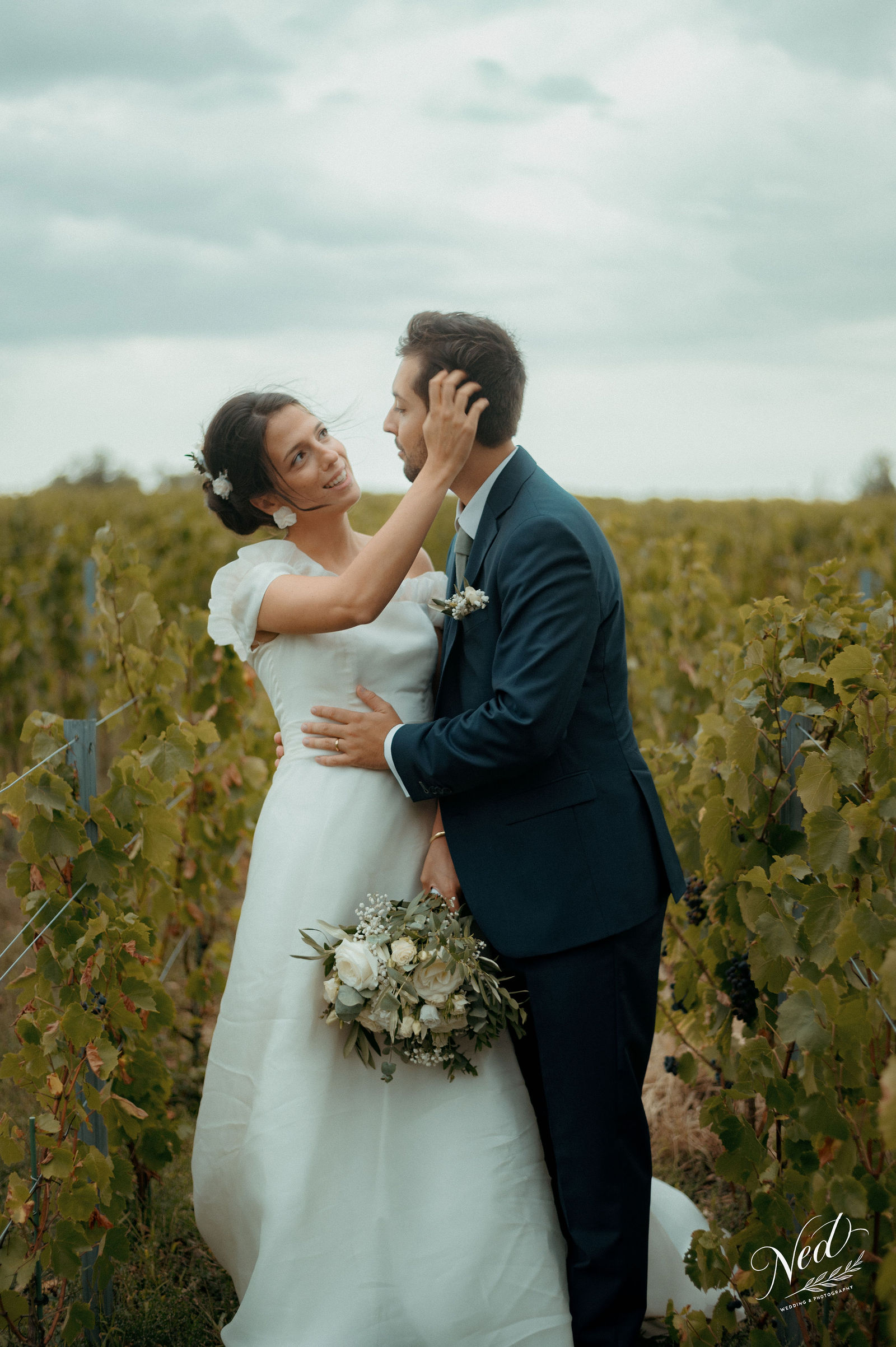 Mariée en robe blanche et marié en costume bleu posent enlacés dans un vignoble, bouquet de fleurs blanches à la main.