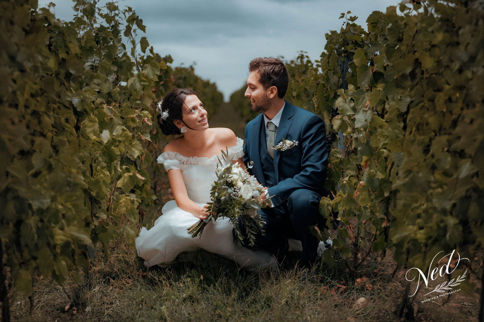 Mariée en robe blanche et marié en costume bleu se regardent entre les rangées de vignes en automne