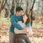 Jeune couple enlacé qui rit sous des arbres en automne, feuilles dorées qui tombent autour d'eux