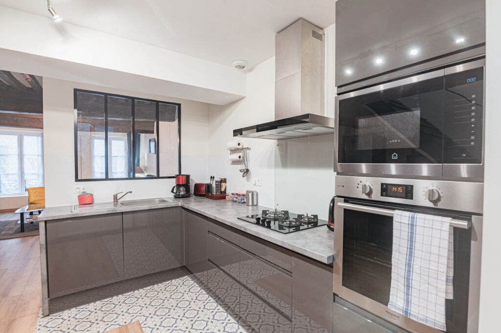 Cuisine contemporaine avec mobilier gris, plan de travail marbre, électroménager inox et fenêtre noire donnant sur séjour lumineux