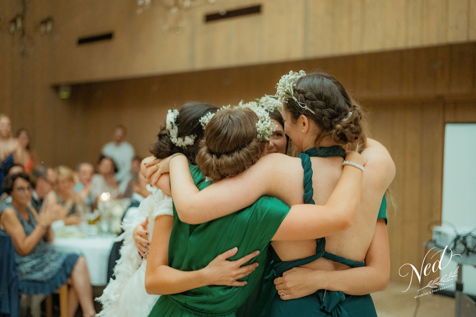 Trois femmes en robes vertes avec fleurs blanches dans les cheveux s'enlacent dans une salle de réception, invités assis en arrière-plan