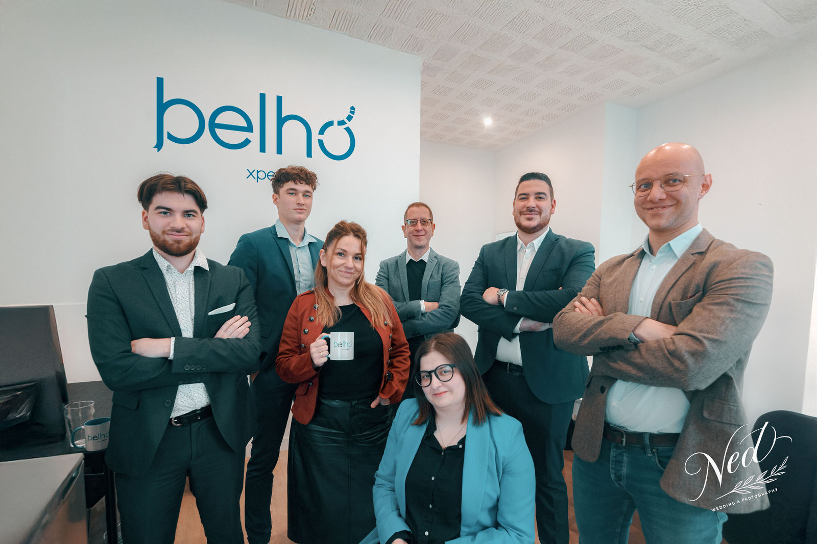 Six femmes et hommes en tenue professionnelle posent ensemble devant un mur blanc avec le logo Belho, dans un bureau moderne.