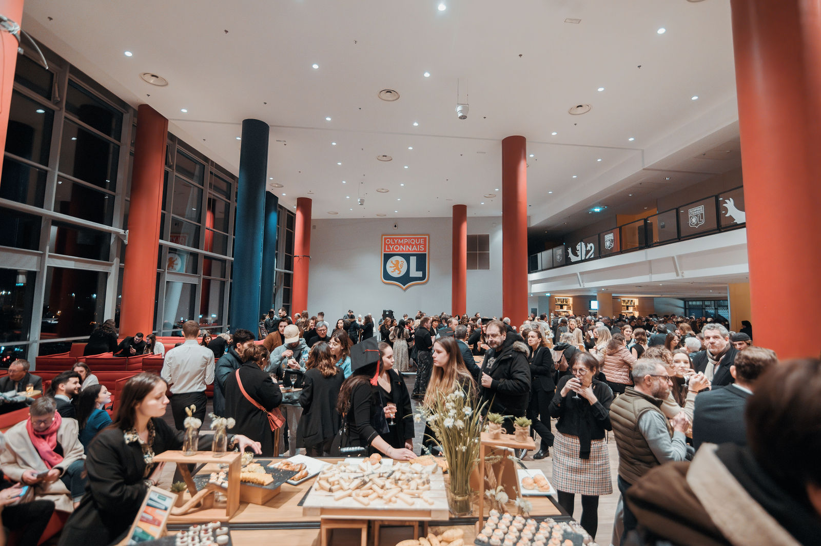 Foule de visiteurs circulant dans un grand hall moderne du stade de l'Olympique Lyonnais, colonnes rouges et bleues, tables de dégustation au premier plan