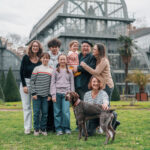 Famille de huit personnes et un chien posent sur la pelouse du Parc de la Tête d'Or à Lyon, serres en verre en arrière-plan