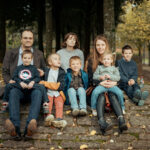 Famille de sept personnes assise sur un banc en pierre dans une allée d'arbres en automne, souriant à la caméra