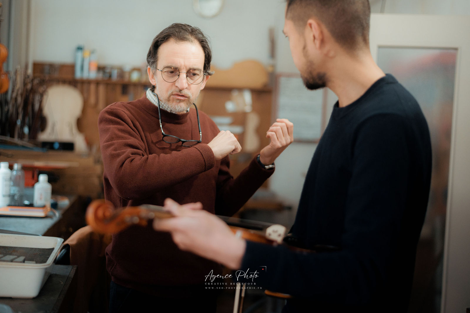 Formateur en pull marron et lunettes explique une technique photo à un jeune participant en noir, dans un atelier avec etabli et materiel photo