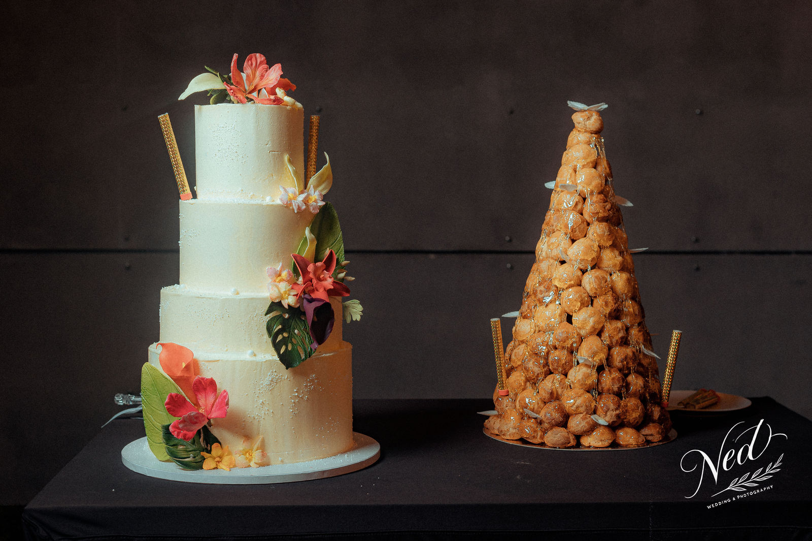 Deux gâteaux de mariage sur socle noir, l'un blanc quatre étages orné de fleurs rouges et bâtons dorés, l'autre pièce montée de choux à la crème caramélisée