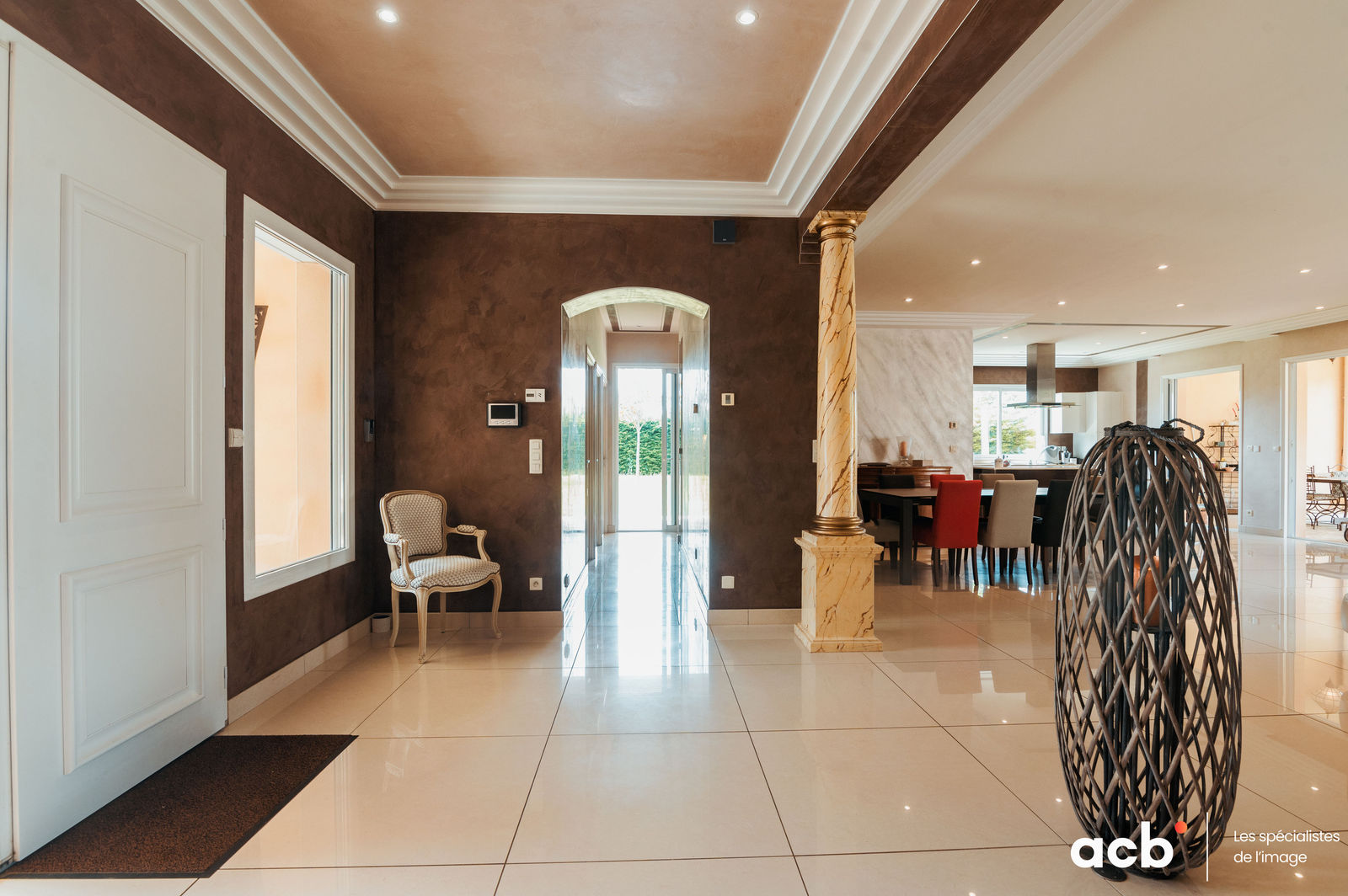 Hall d'entrée avec murs marron, carrelage beige brillant, colonne dorée, porte-parapluies noir et accès vers salon lumineux