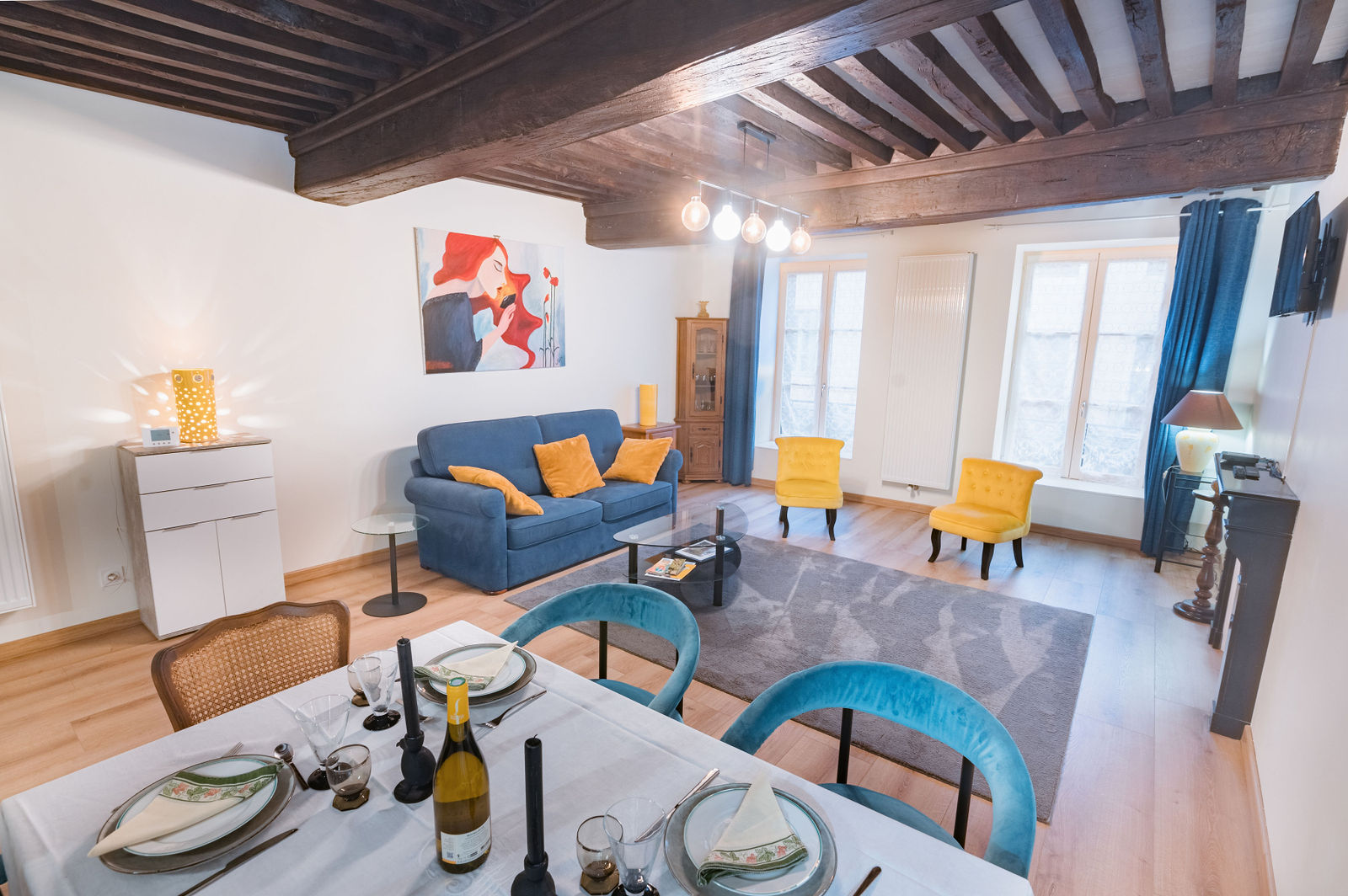 Loft spacieux avec poutre apparente, canapé bleu, fauteuils jaunes, table à manger en premier plan, lumière naturelle par les portes vitrées