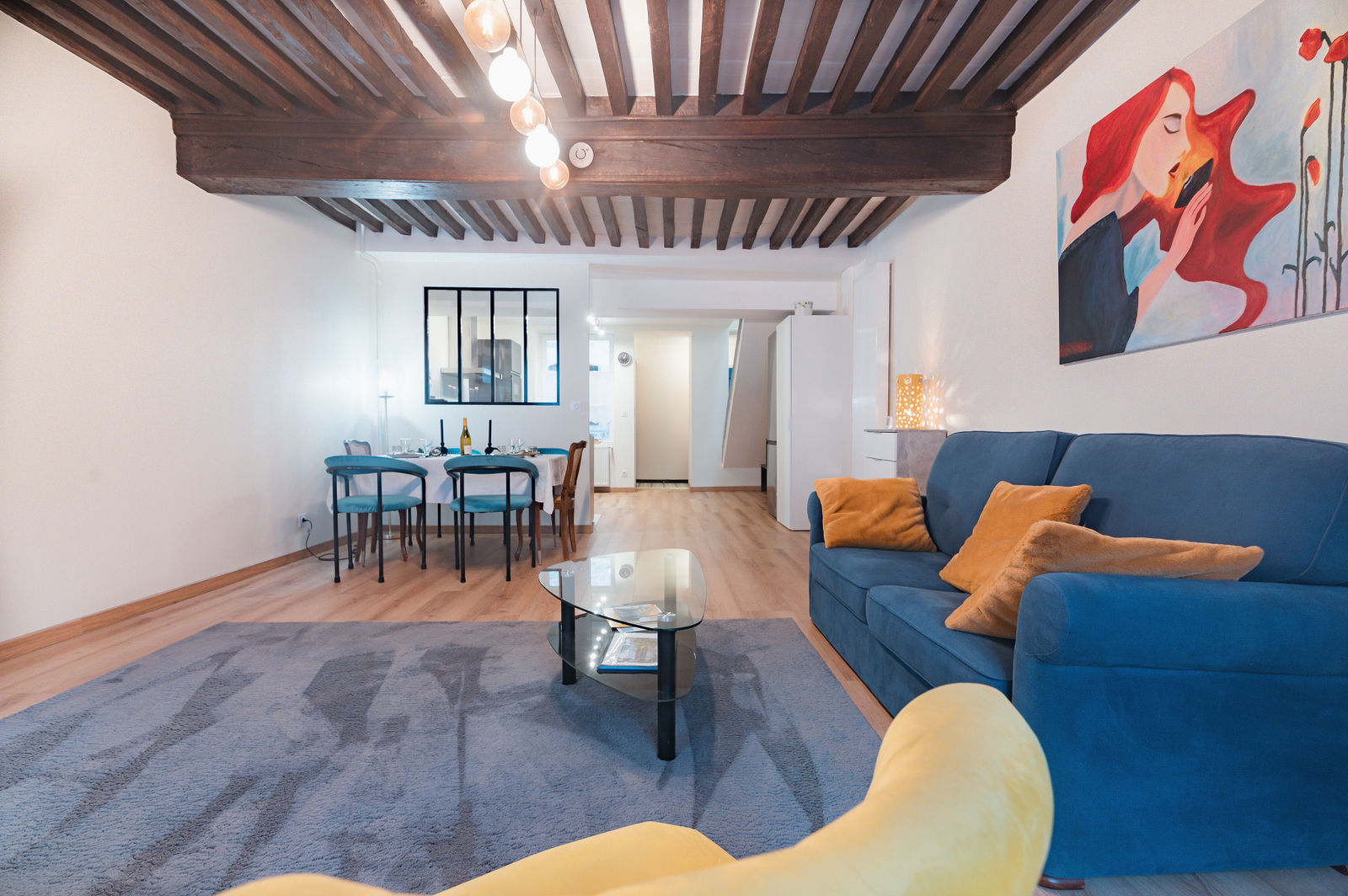 Salon spacieux avec canapé bleu, table basse en verre, poutres en bois au plafond et chaises bleues près de la salle à manger